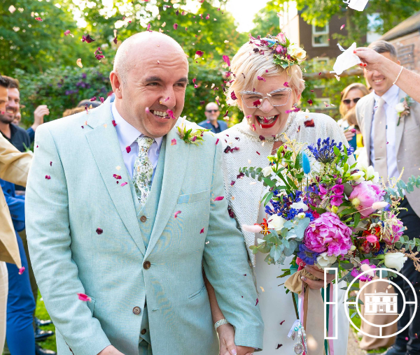 Bride and groom walking through garden as guests throw confetti.