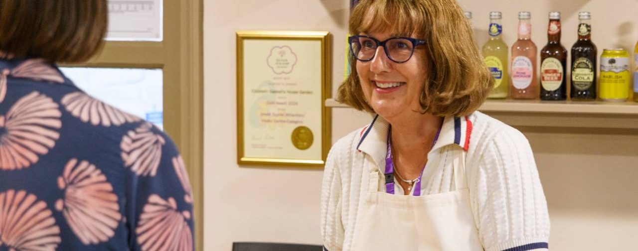 A volunteer with brown hair in a bob wearing glasses smiling at a visitor in a tea room.