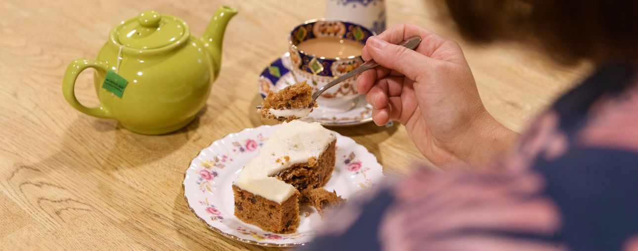 A green teapot, blue and white cup and saucer with tea in it and a small plate with a piece of cake on it. A hand is holding a fork with a piece of cake on it.