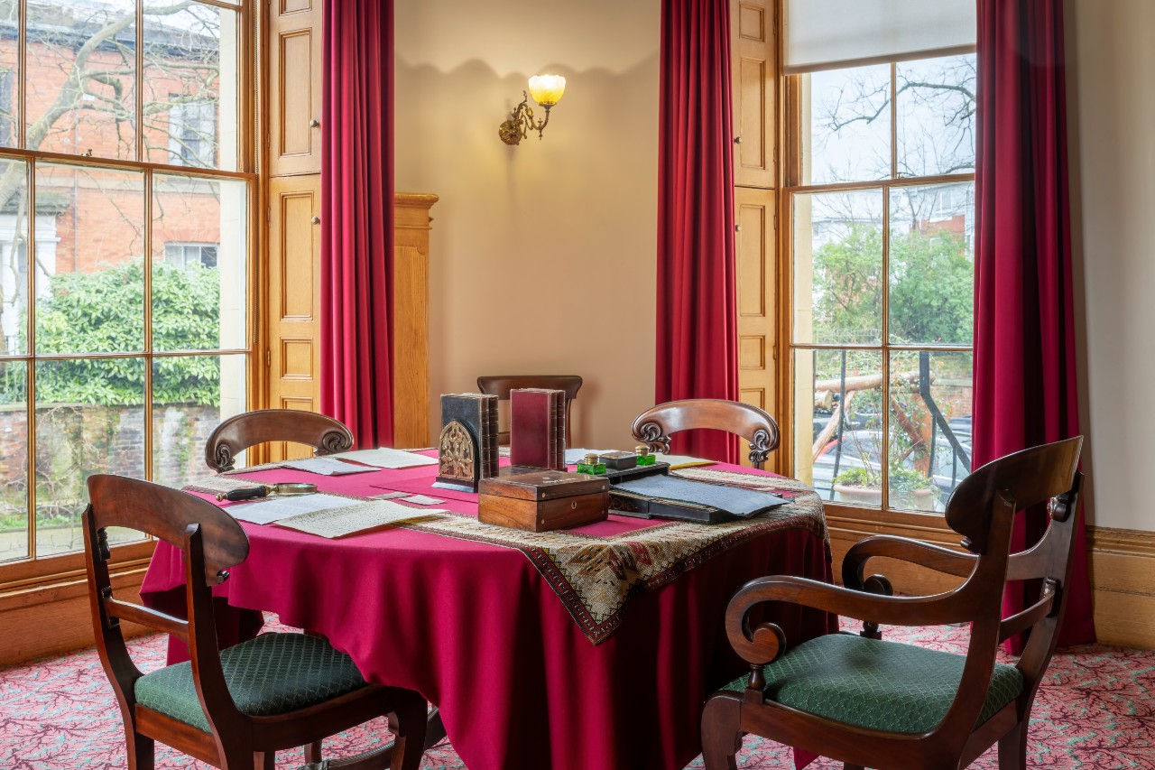 The round writing table in the Elizabeth Gaskell's House Dining Room, with a red tablecloth, surrounded by dining chairs with green seats. The table has books and manuscripts on it, and is surrounded on all sides by large, sunny windows.