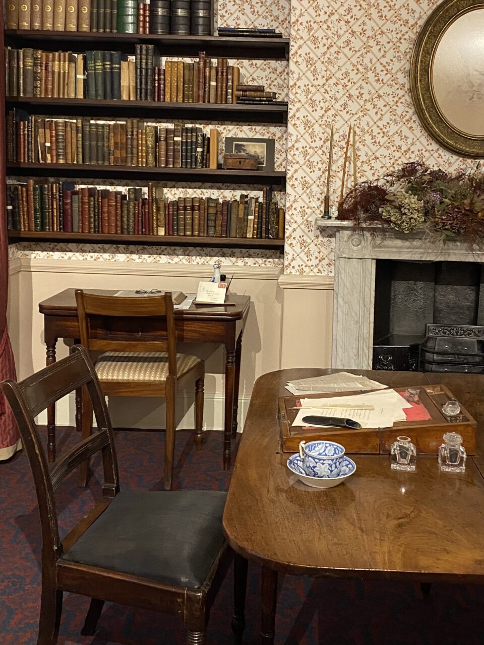 A room at the Bronte Parsonage. There is a brown table and chair, and behind it is a fireplace and bookshelves.