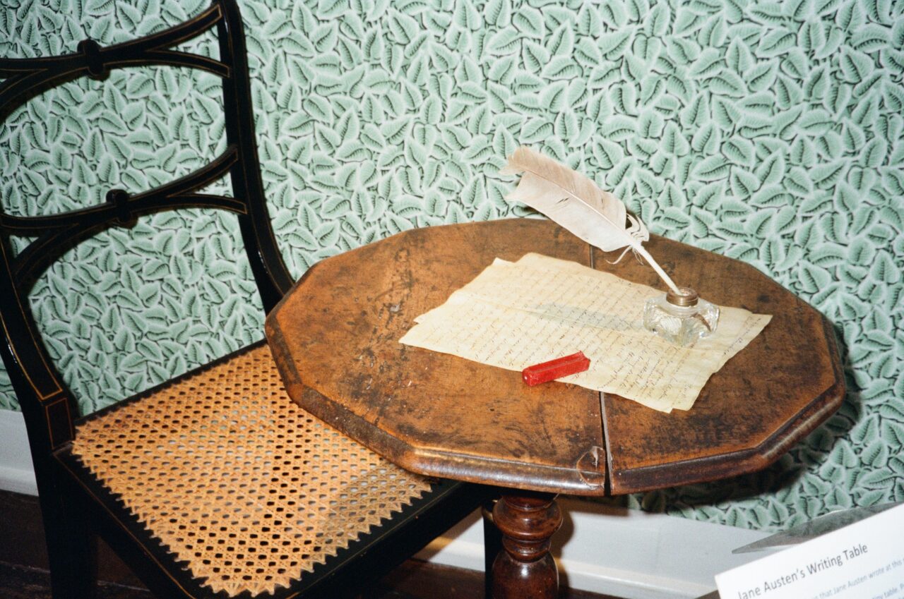 Jane Austen's writing desk, a small, brown, octagonal table, with a piece of paper and a quill on top of it. There is a dark chair behind the table and the wallpaper behind it is bright turquoise. In the corner you can see a small plaque which states this is Jane Austen's writing desk.
